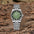 Reloj Calma con esfera verde y brazalete de acero, en posición vertical sobre un fondo de madera natural